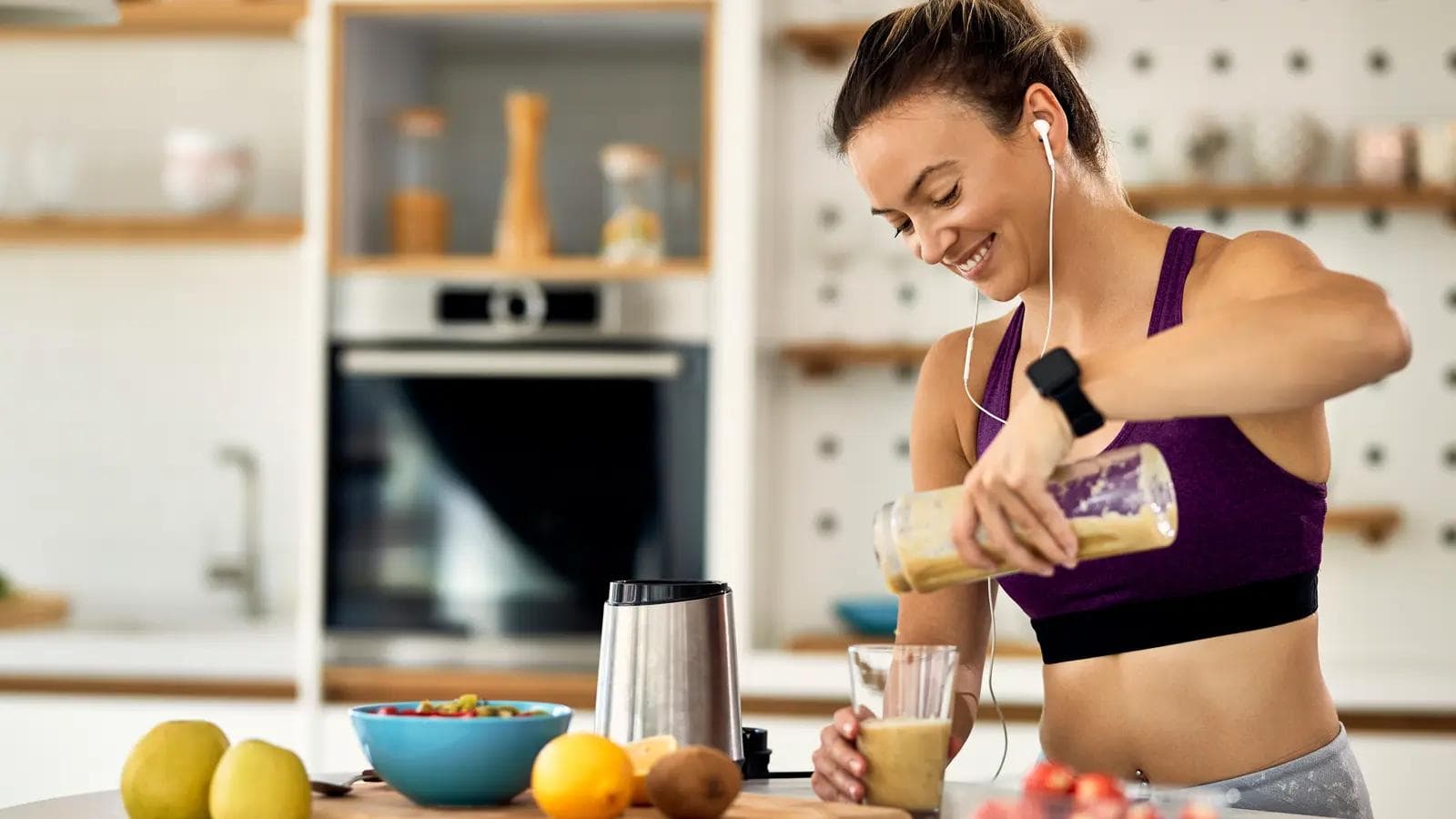 Nutrition for Beginners: The 5 'Building Blocks' for a Fitter, Stronger You Smiling woman in sportswear pouring a healthy smoothie from a blender bottle into a glass in a bright kitchen.